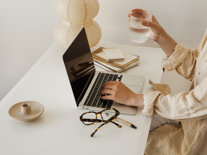 A person working at a white desk with a laptop, notebooks, a pair of glasses, a glass of water, a ceramic vase, and a paper lantern.