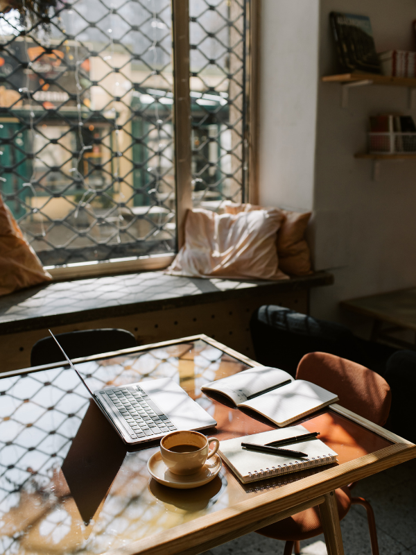 Cozy workspace with a wooden table featuring a laptop, a cup of coffee on a saucer, an open notebook, and a pen. The table is near a window with a metal grid, letting in natural light, with soft pillows on the window seat and a few shelves on the wall.