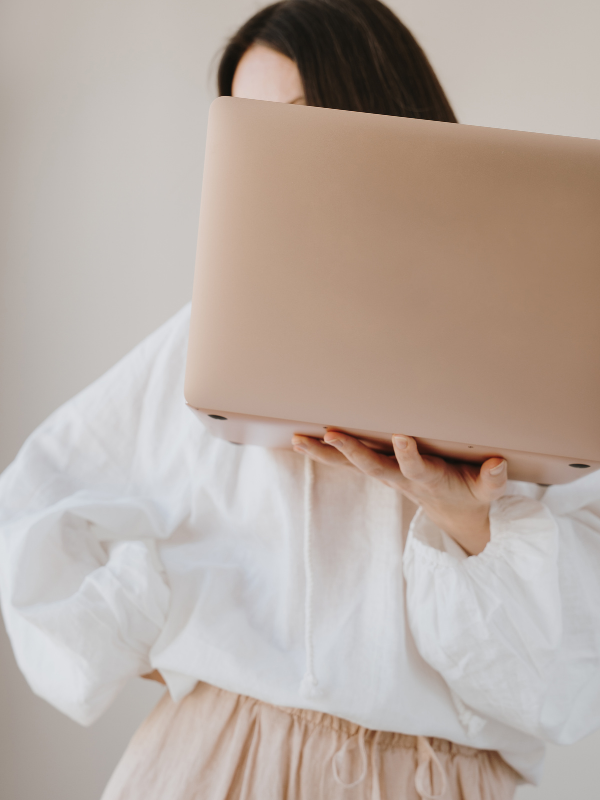 Person holding a closed beige laptop in front of their face, wearing a white jacket and beige pants.