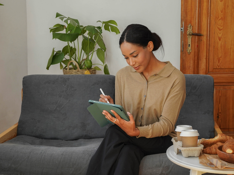 Woman sitting on a gray sofa, writing on a tablet with a stylus, with coffee cups and pastries on a table nearby, in a room with a potted plant and a wooden door.