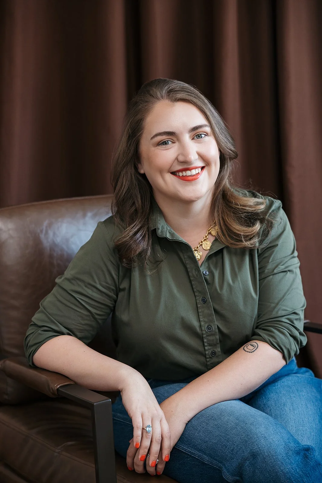 A woman with shoulder-length brown hair, wearing a dark green button-up shirt, gold necklace, and red lipstick, sitting on a brown leather chair in front of brown curtains, smiling at the camera.