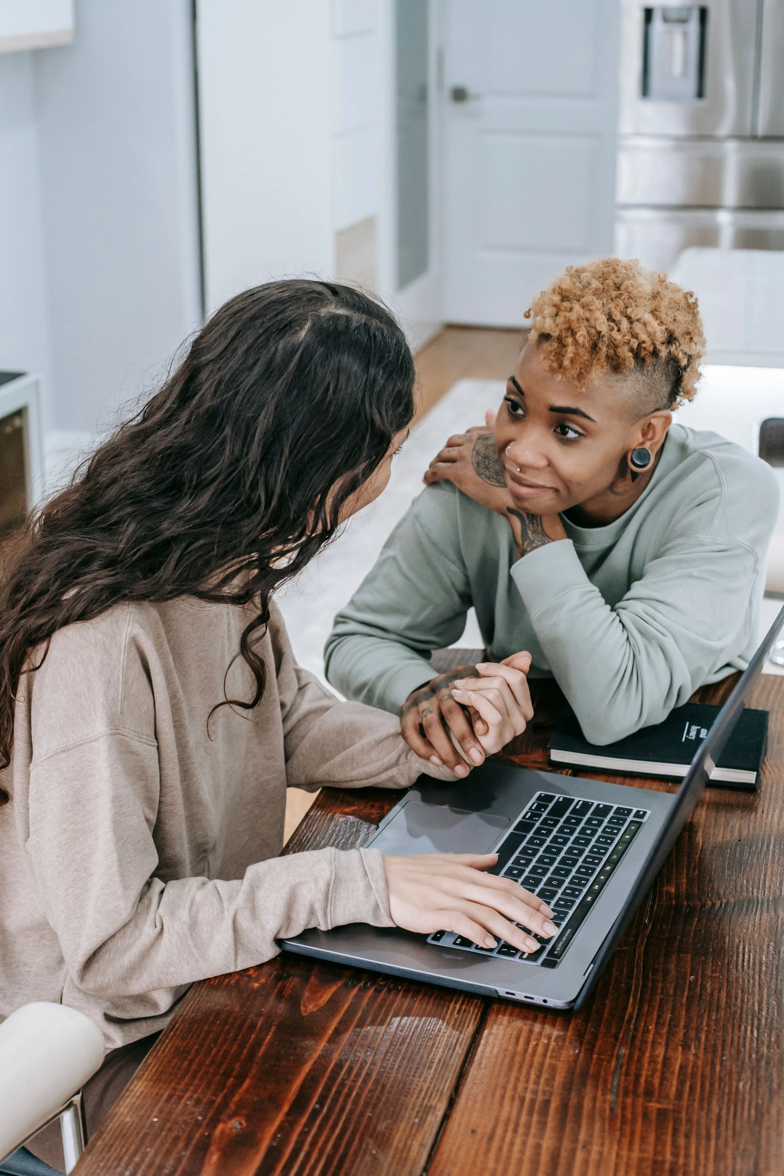 Two women sitting at a wooden table, holding hands, with a laptop in front of them. One woman has long dark hair, and the other has short, curly blonde hair and visible tattoos.