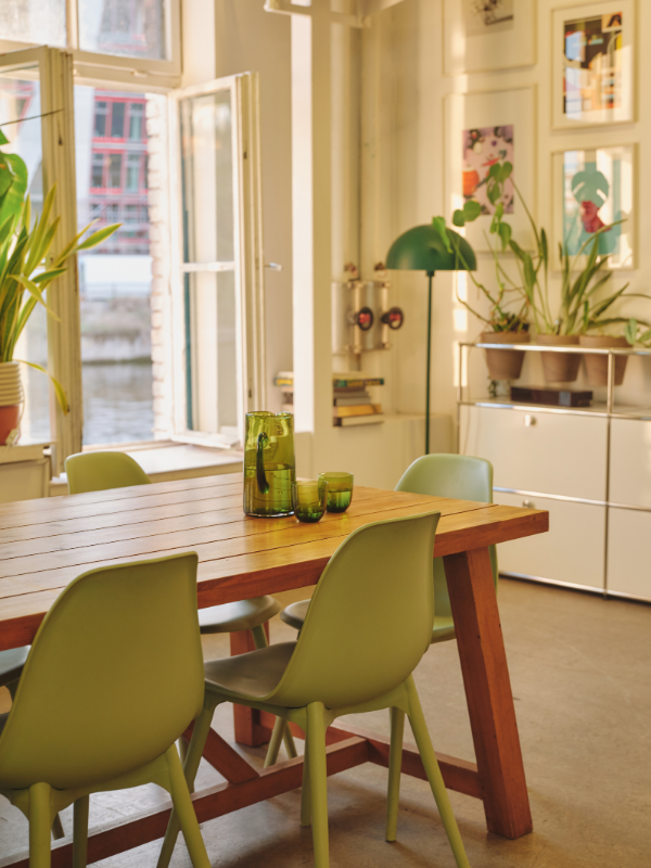 Bright dining room with open window, wooden table with green glassware, and green chairs, decorated with potted plants and artwork on the walls.