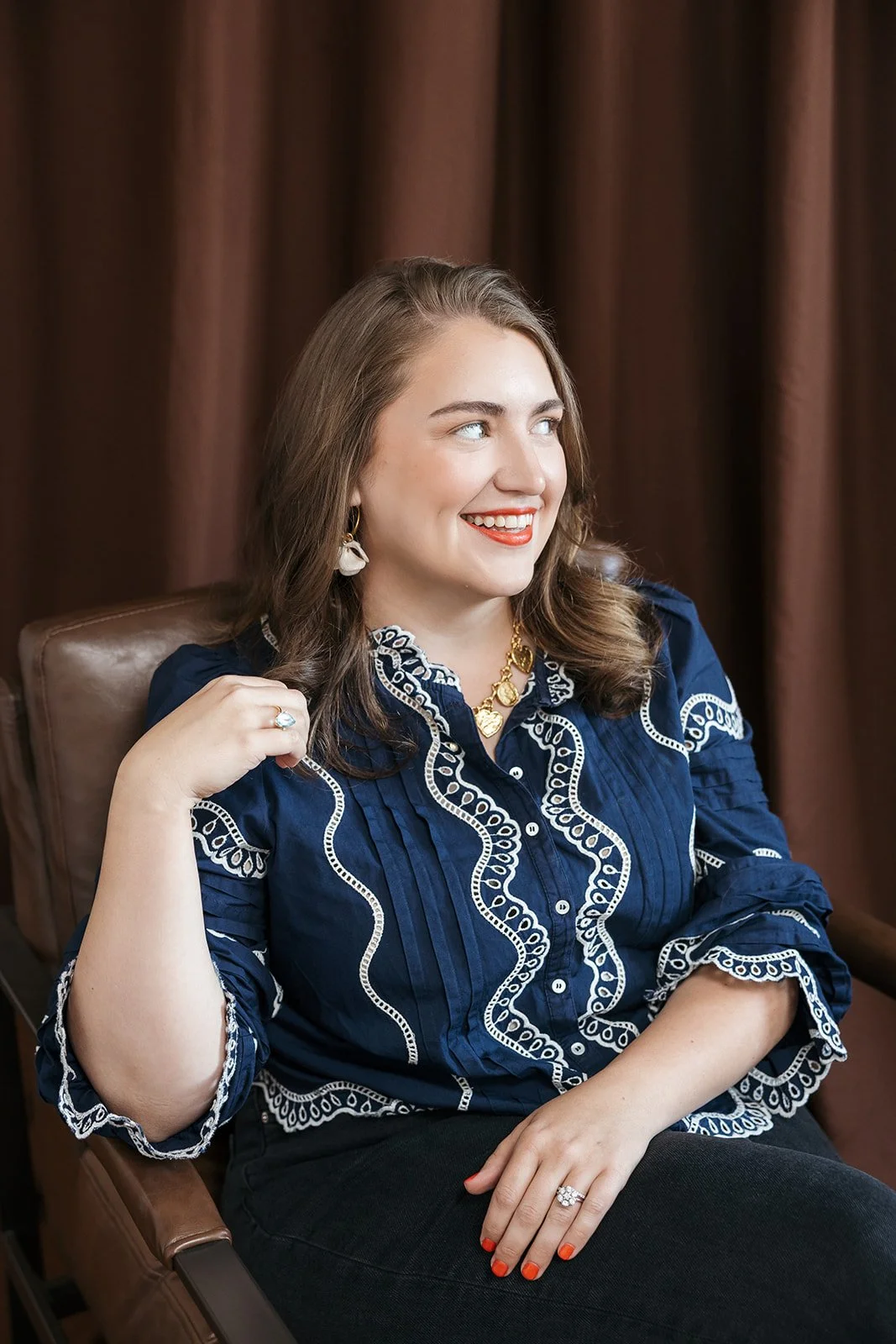 A woman sitting in a chair with brown curtains in the background, smiling and looking to her right. She is wearing a blue embroidered blouse, white earrings, gold necklace, a ring, and has painted red nails.