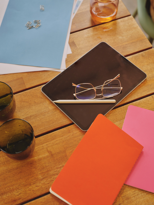 A wooden table holding a tablet with glasses and a stylus, a pair of sunglasses on the tablet, pink and orange notebooks, two dark glass cups, a glass of iced tea, and some papers and clips.