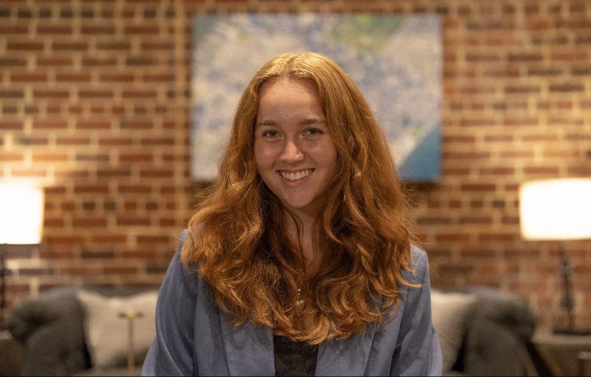 A woman with long curly red hair smiling in a cozy living room with brick walls, a map, and lamps.