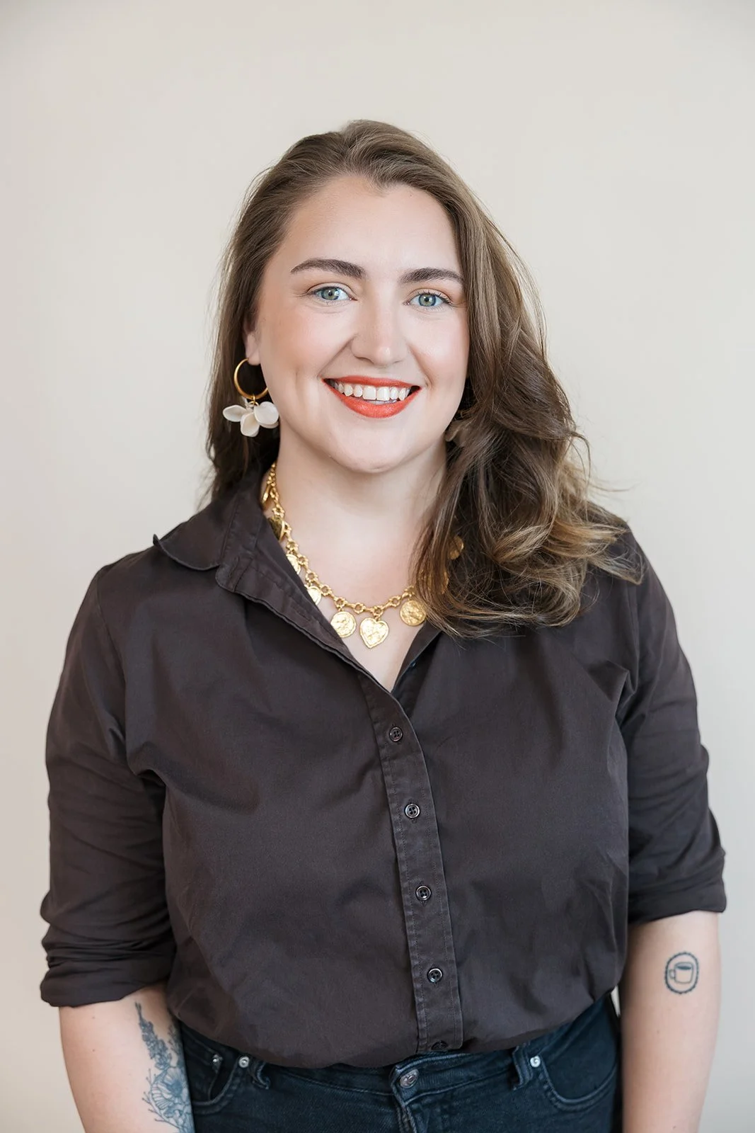 A woman with long wavy brown hair, wearing a dark button-up shirt, gold jewelry, and hoop earrings, smiling against a plain light background.