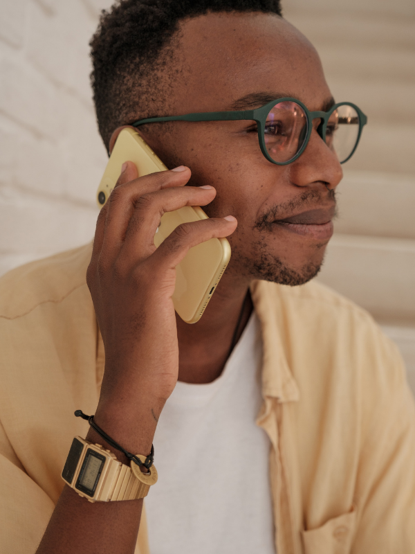 Man with glasses talking on a yellow smartphone, wearing a beige shirt, white t-shirt, bracelet, and watch.