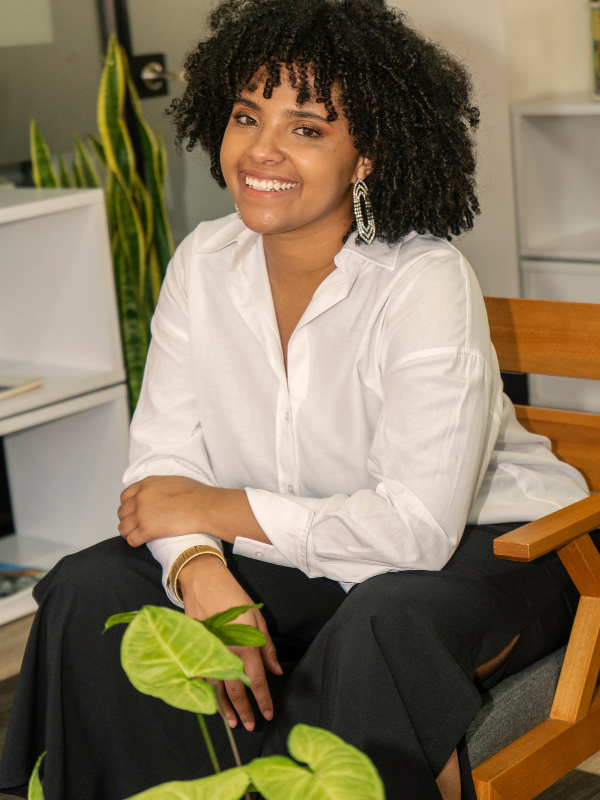 Young woman with curly hair smiling, sitting on a chair in an office with plants and shelves in the background.