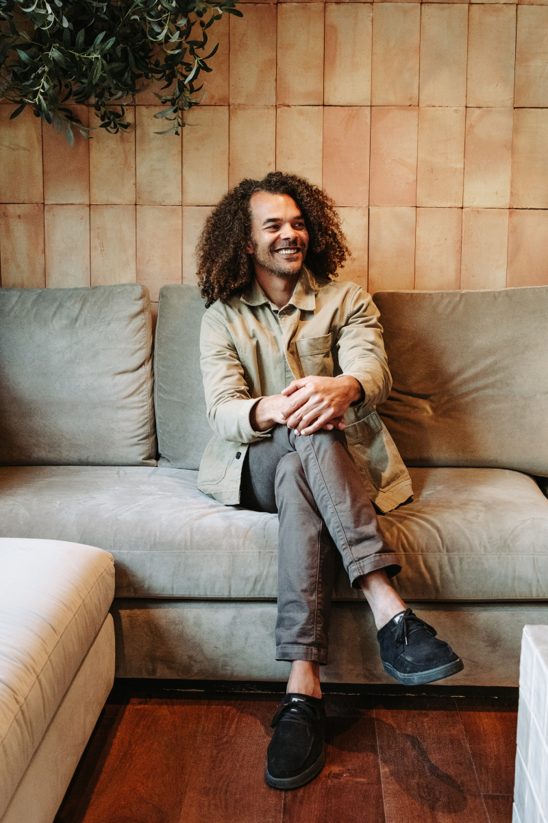 A man sitting on a beige couch in a room with wooden-paneled walls and a green plant in the corner, smiling and looking to the side.