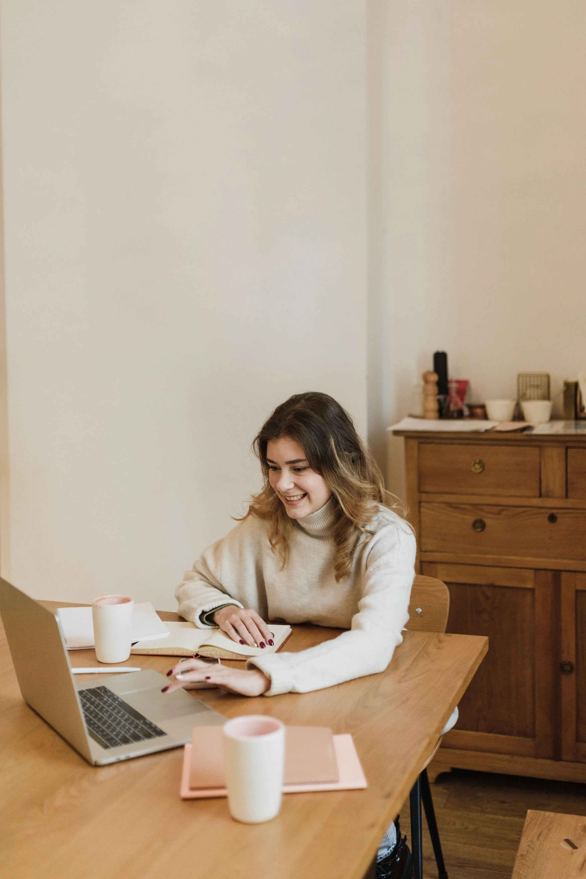 A young woman with long, wavy brown hair sitting at a wooden table using a laptop, with open notebooks and a white mug in front of her in a cozy, well-lit room with wooden furniture.