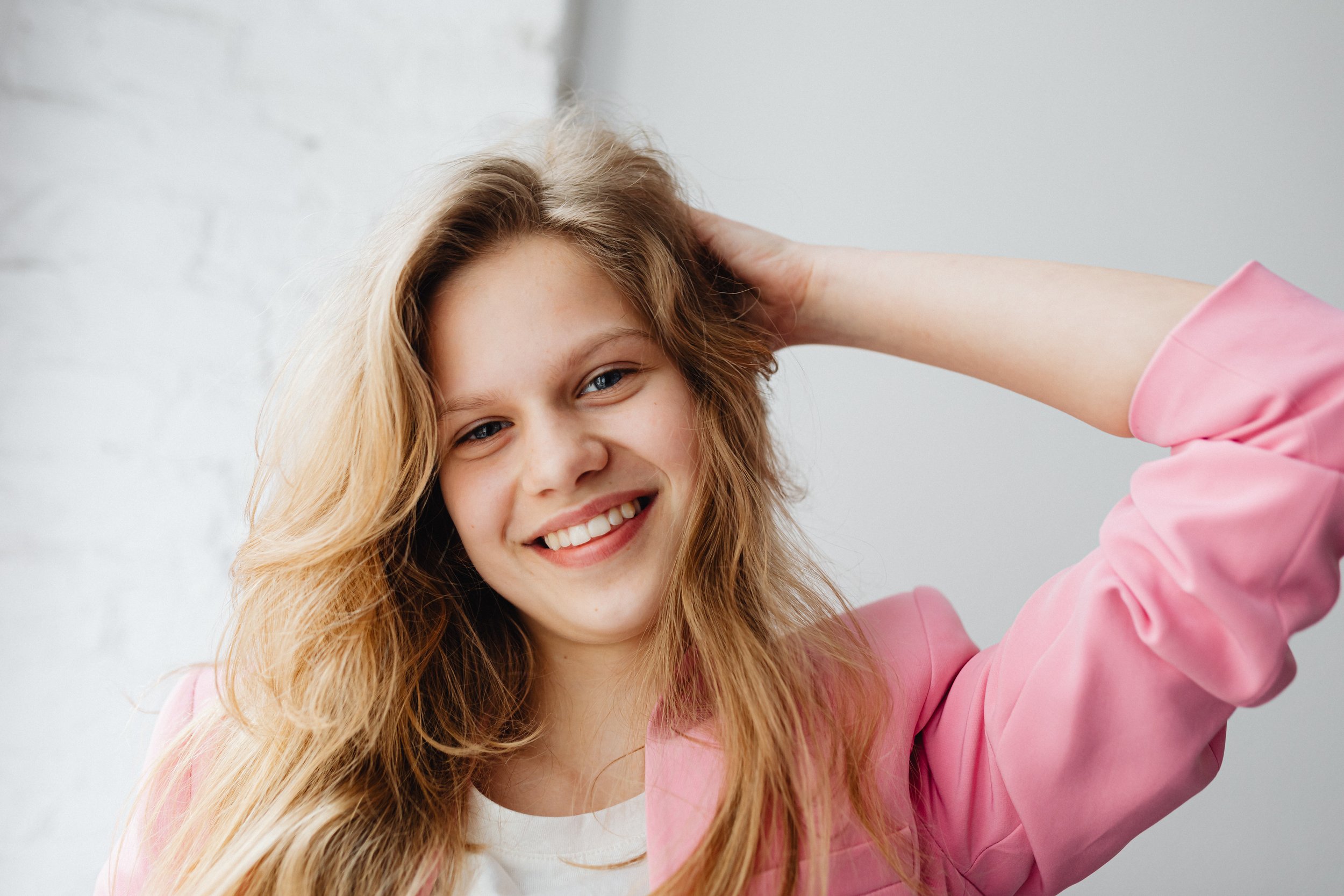 A young woman with long, wavy blonde hair smiling and holding her head with her right hand, wearing a pink jacket and a white top, standing against a light-colored background.