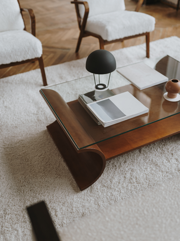 A glass coffee table with a wooden base, placed on a white shag rug in a living room. The table has a black desk lamp, a stack of magazines, some white books, and a small brown vase.