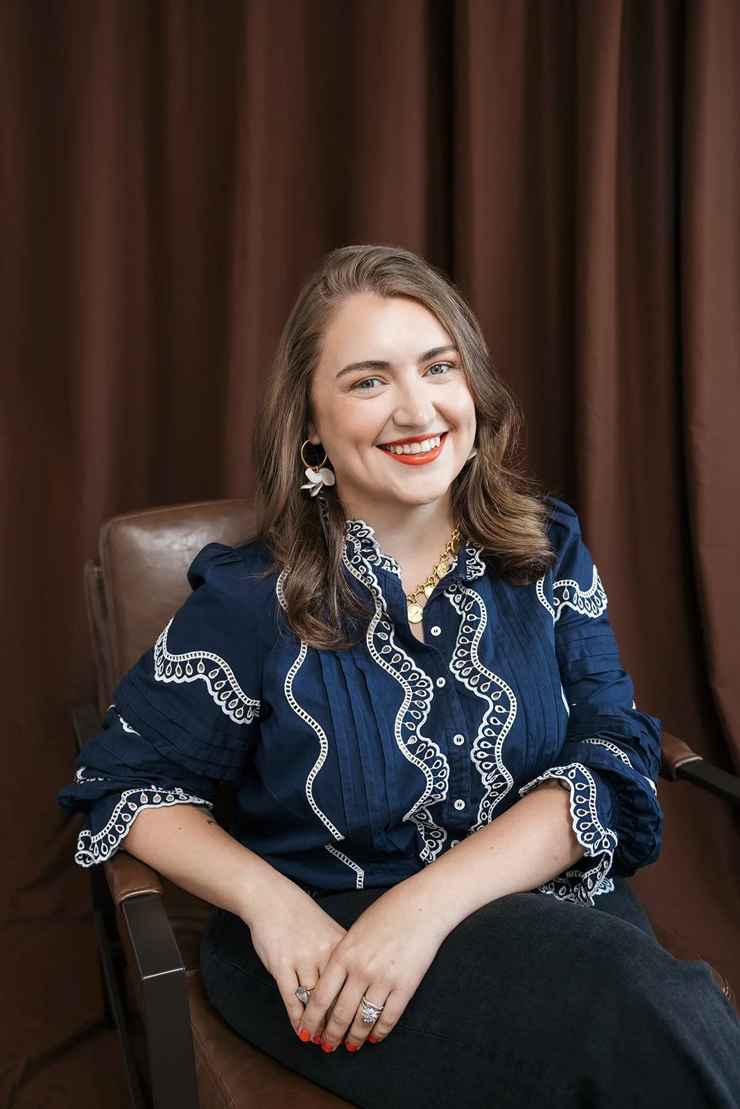 A woman with long brown hair, wearing a navy blue blouse with white embroidery, gold jewelry, and earrings, sitting on a brown chair in front of brown curtains, smiling at the camera.