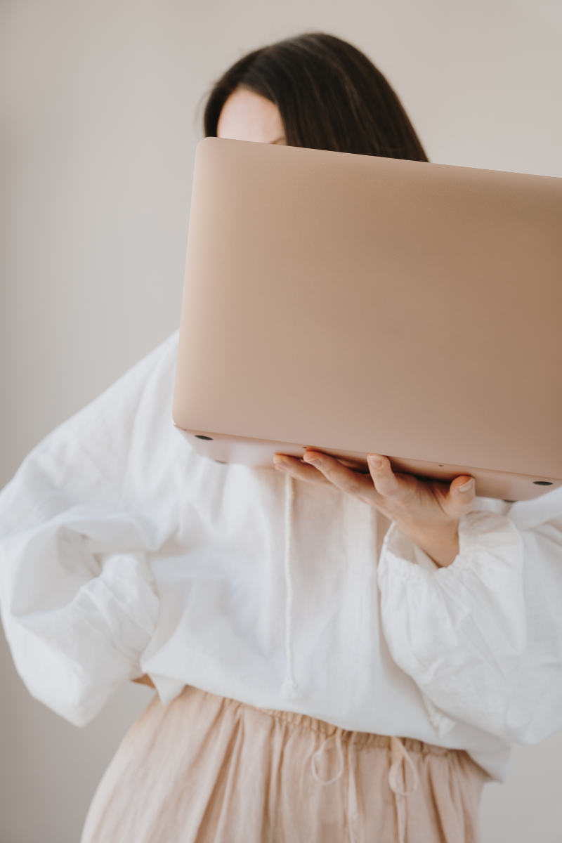 A woman holding a beige laptop, partially covering her face, wearing a white blouse and beige pants.