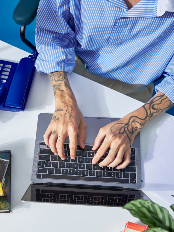 Person with tattoos working on a laptop at a desk, with a blue shirt and office supplies around.