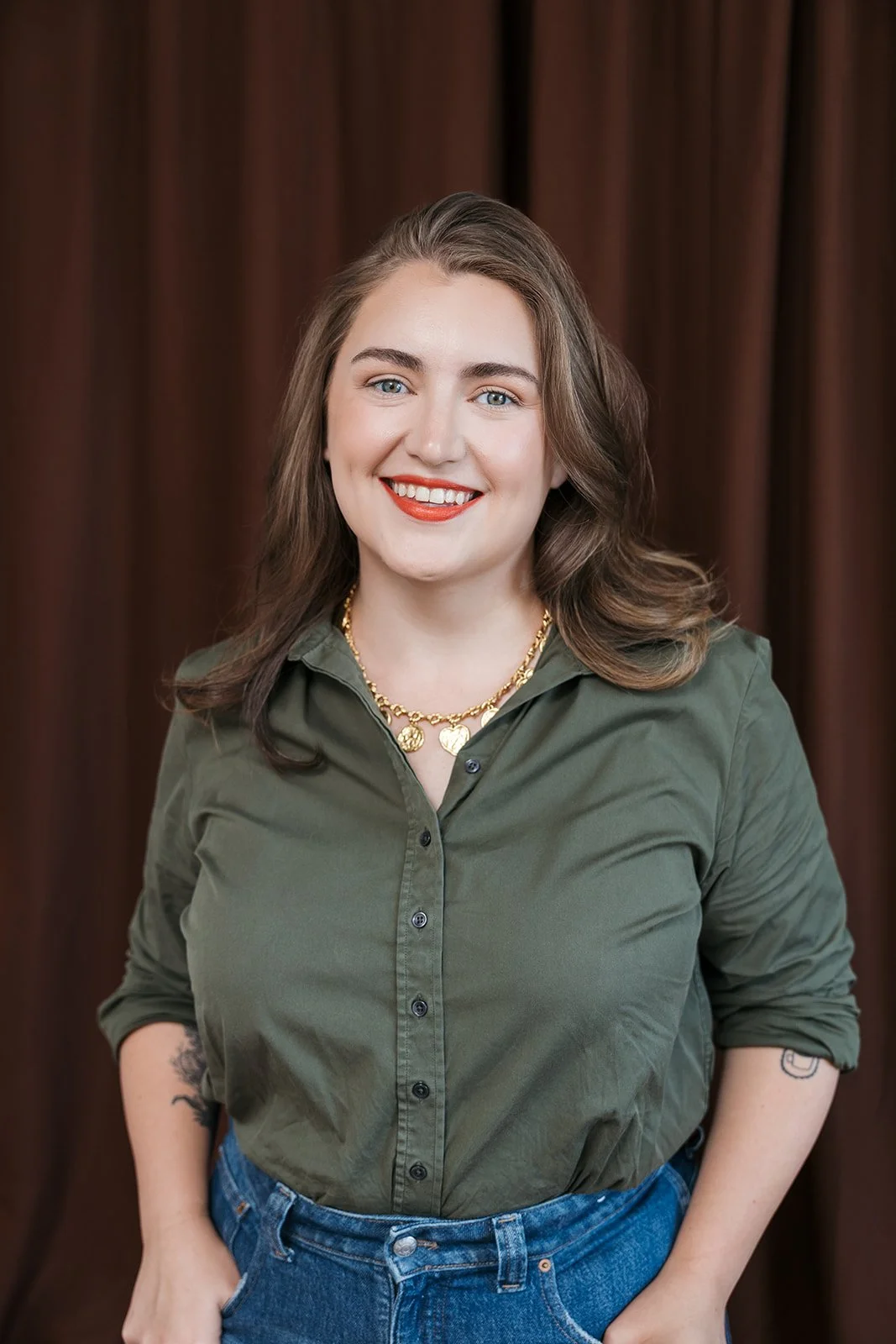 A woman with shoulder-length wavy brown hair, wearing a green button-up shirt and blue jeans, smiling with red lipstick, gold chain necklace, and tattoos on her arms, standing in front of brown curtains.