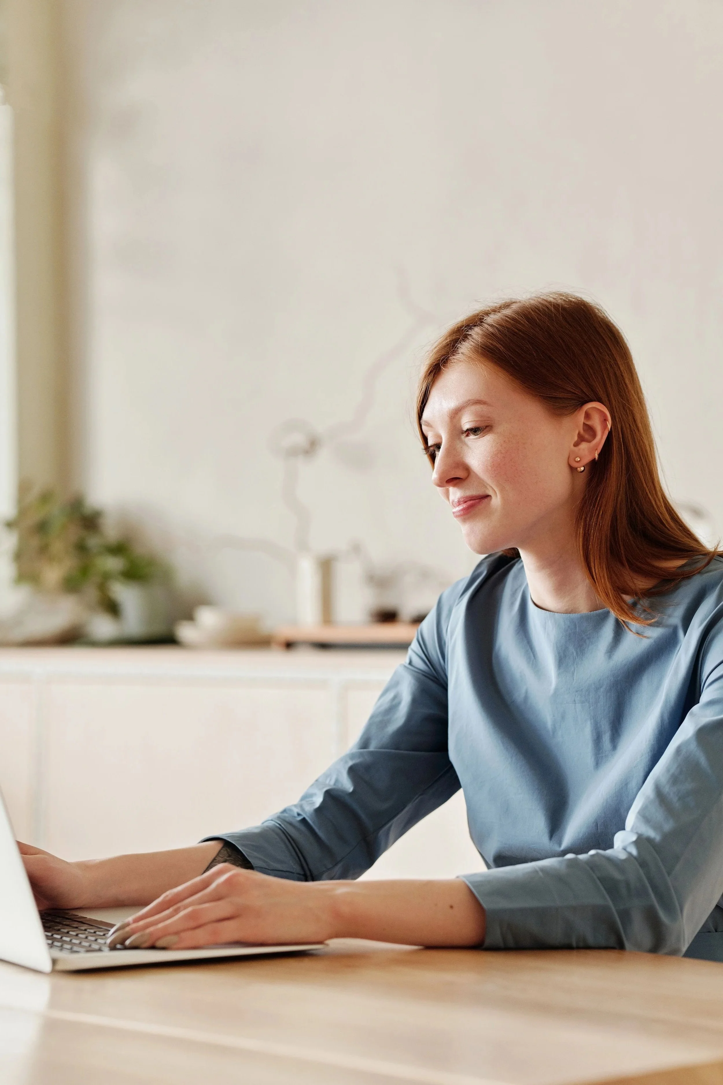 A woman with red hair and earrings is working on a laptop at a desk in a bright room.