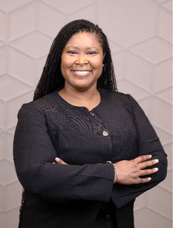 A smiling woman with long braided hair wearing a black blazer, standing with arms crossed in front of a light gray geometric wall.