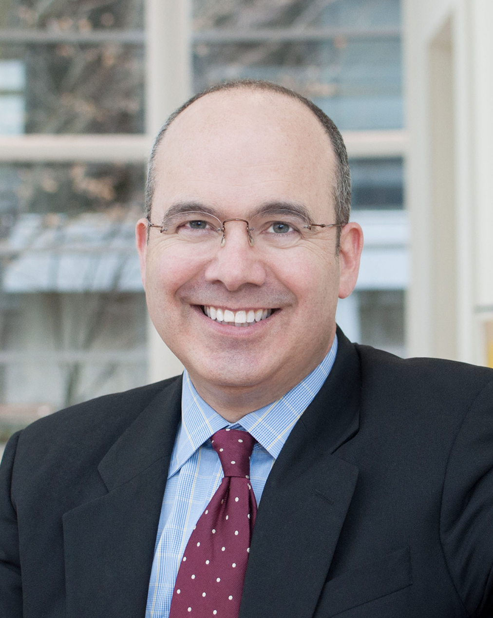 A smiling man in a suit with glasses, wearing a red polka dot tie, sitting indoors with large windows in the background.
