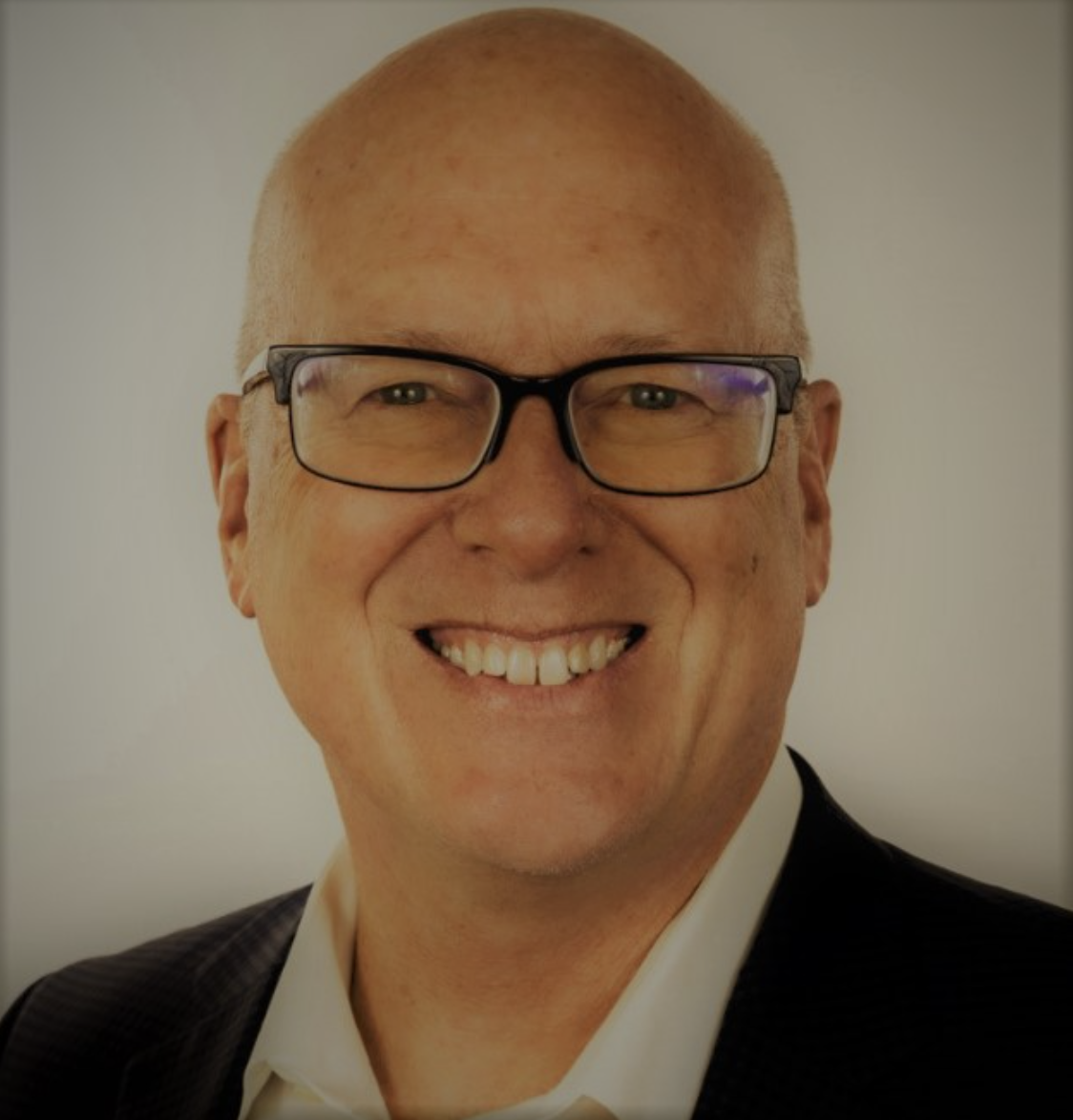 A smiling man with glasses, a bald head, wearing a suit, against a plain background.