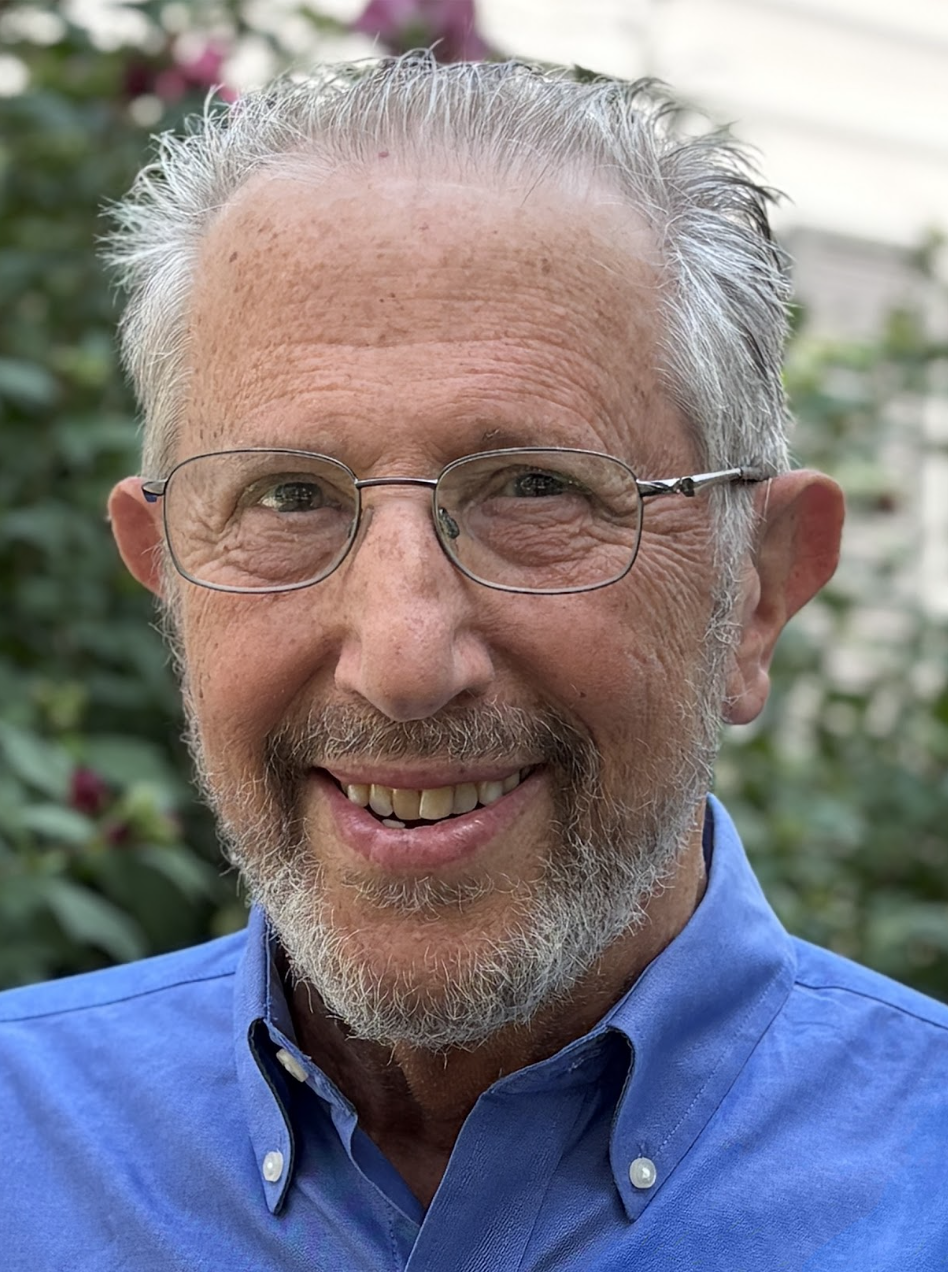 A smiling older man with gray hair, glasses, and a beard, wearing a blue collared shirt, outdoors with greenery and some pink flowers in the background.