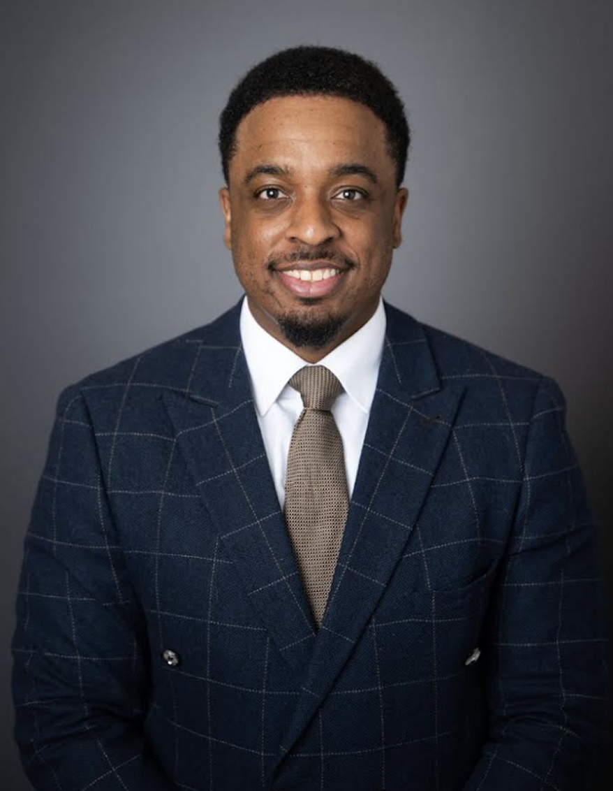 Professional headshot of a smiling Black man in a navy checkered suit, white shirt, and beige tie against a gray background.