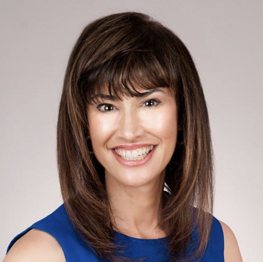 A woman with shoulder-length brown hair, wearing a sleeveless royal blue top, smiling with teeth showing, against a light gray background.