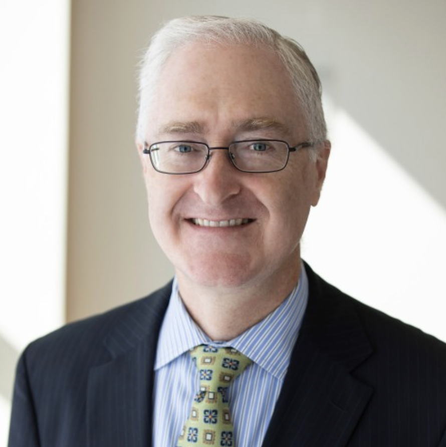 Portrait of a middle-aged man with white hair, wearing glasses, a dark suit, a light blue striped shirt, and a patterned tie, smiling in an indoor setting.