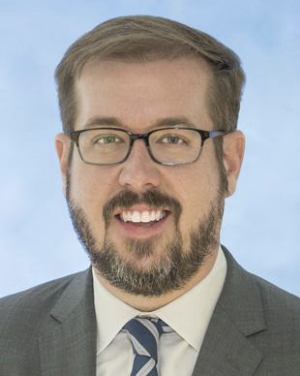 A man with glasses, a beard, and mustache wearing a suit and tie, smiling against a light blue background.