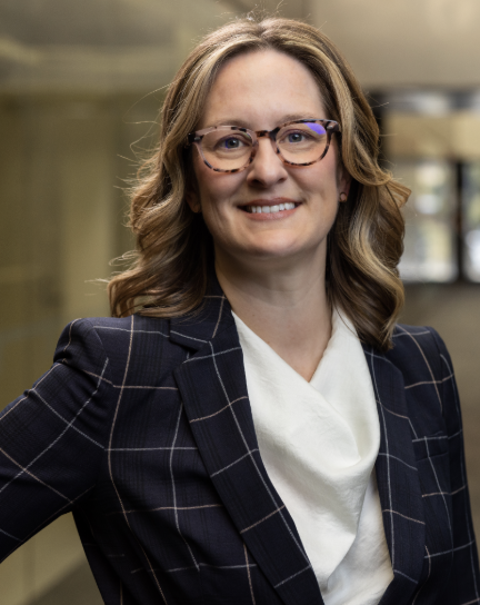 A woman with shoulder-length wavy hair wearing glasses, a dark plaid blazer, and a white shirt, smiling in an indoor setting.