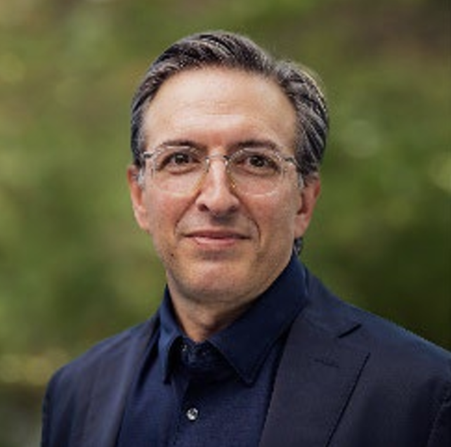 Portrait of a man with glasses, wearing a navy suit and dark shirt, outdoors with a blurred green background.