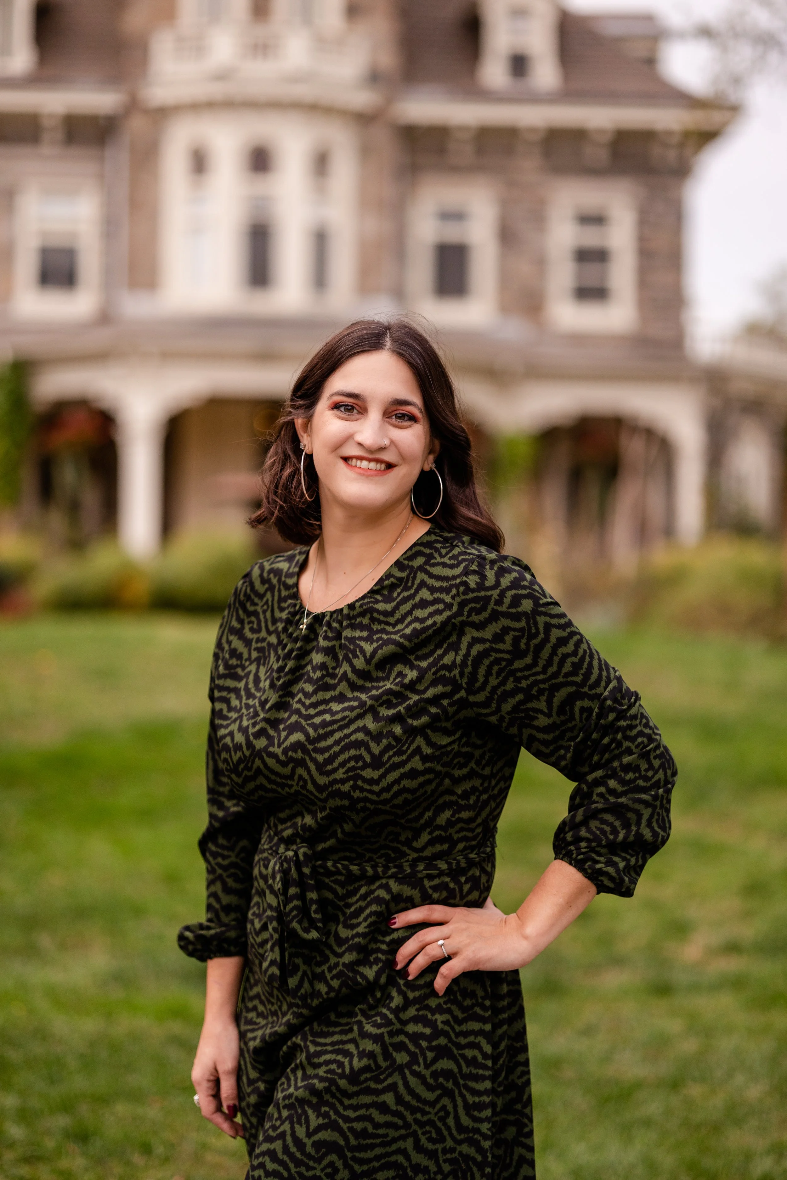 A woman with dark brown hair, wearing a black and green patterned dress, standing outdoors in front of a large house with a porch and multiple windows. She is smiling with her right hand on her hip and wearing hoop earrings and a necklace.