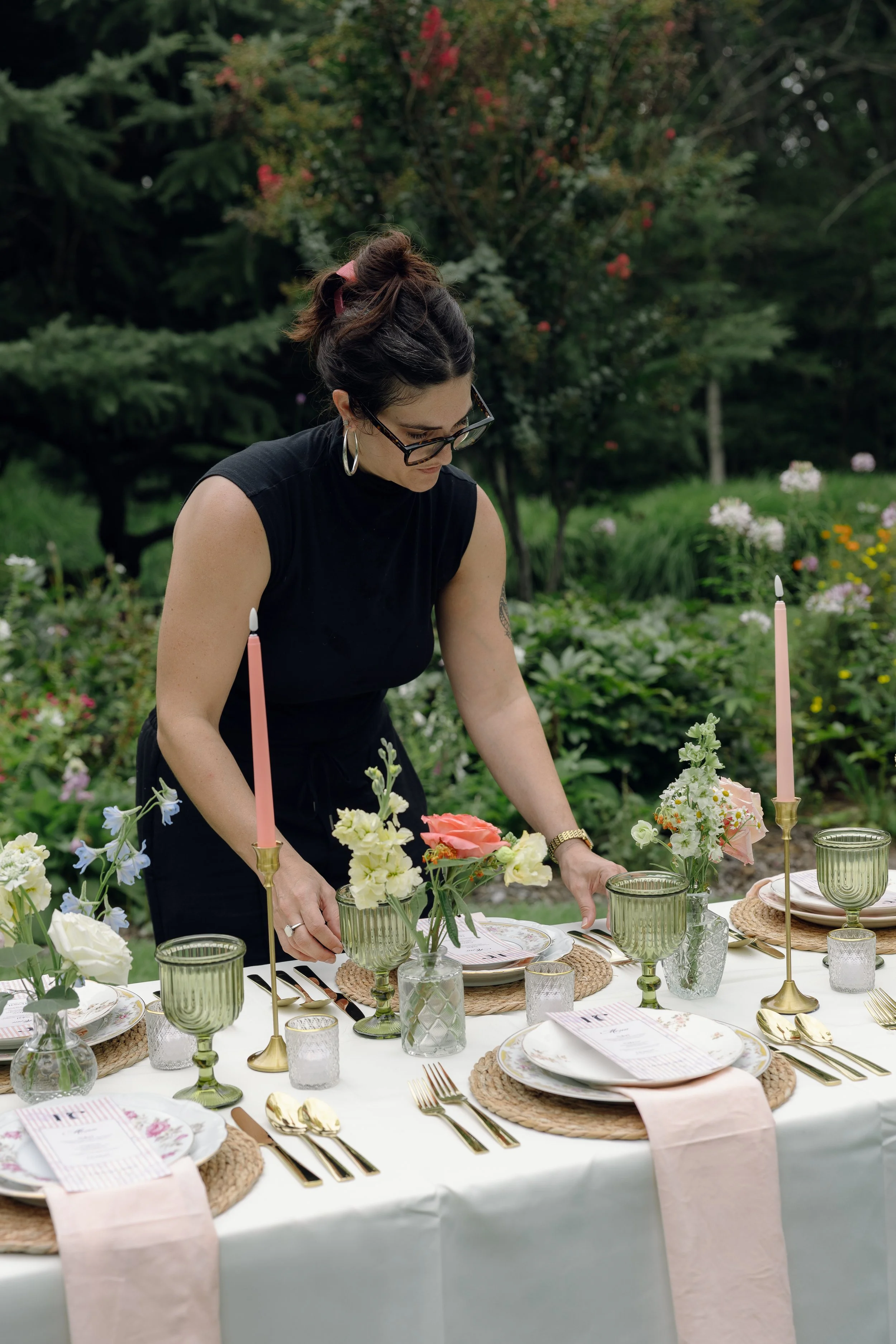 A woman with dark hair in a bun, wearing glasses and a sleeveless black top, arranges a floral centerpiece at an outdoor table set for a formal event in a garden with trees and flowers.