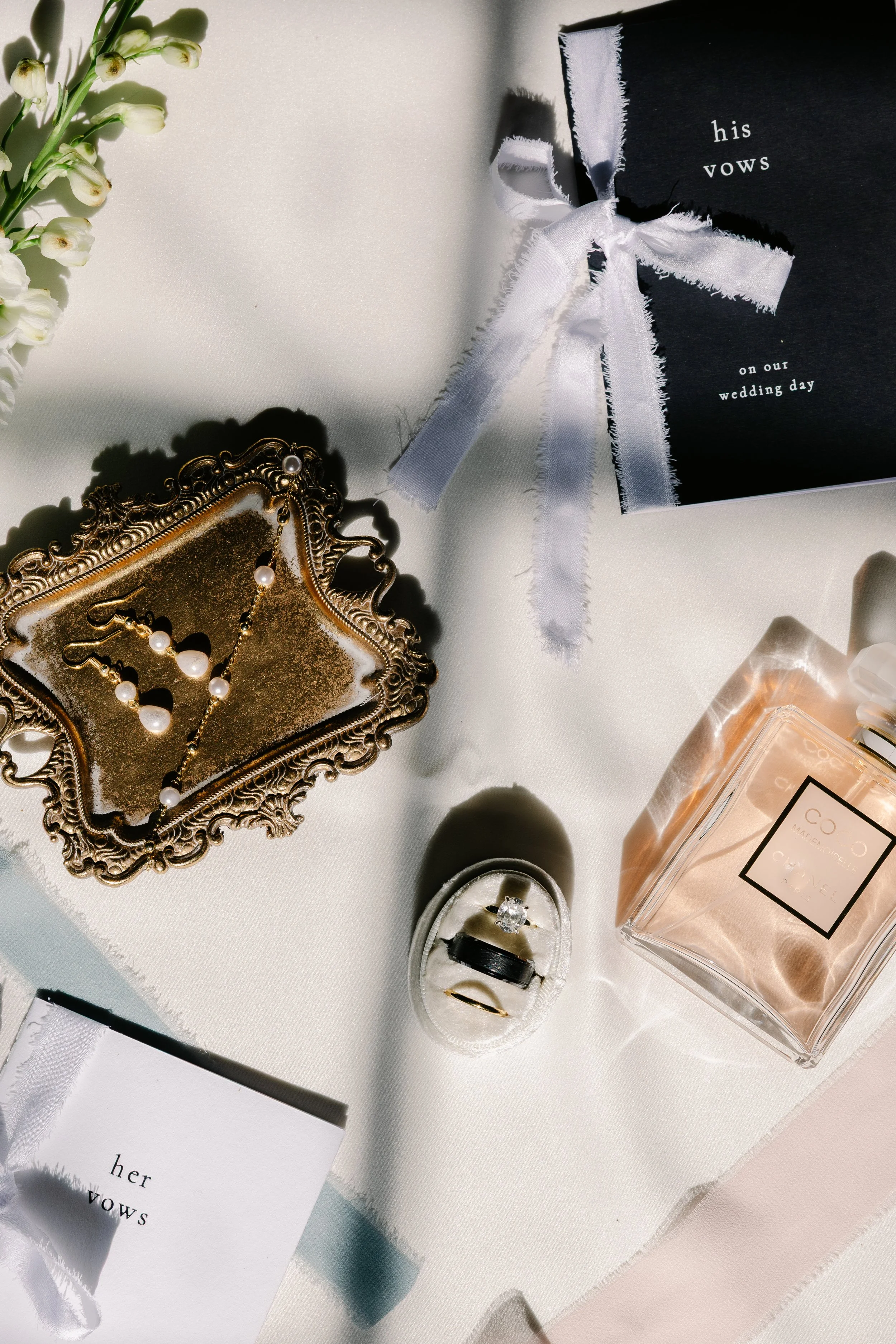 A wedding gift box with a white ribbon, a jewelry dish with pearl earrings, floral accents, wedding vows card, a perfume bottle, and rings in a small velvet box on a white surface.