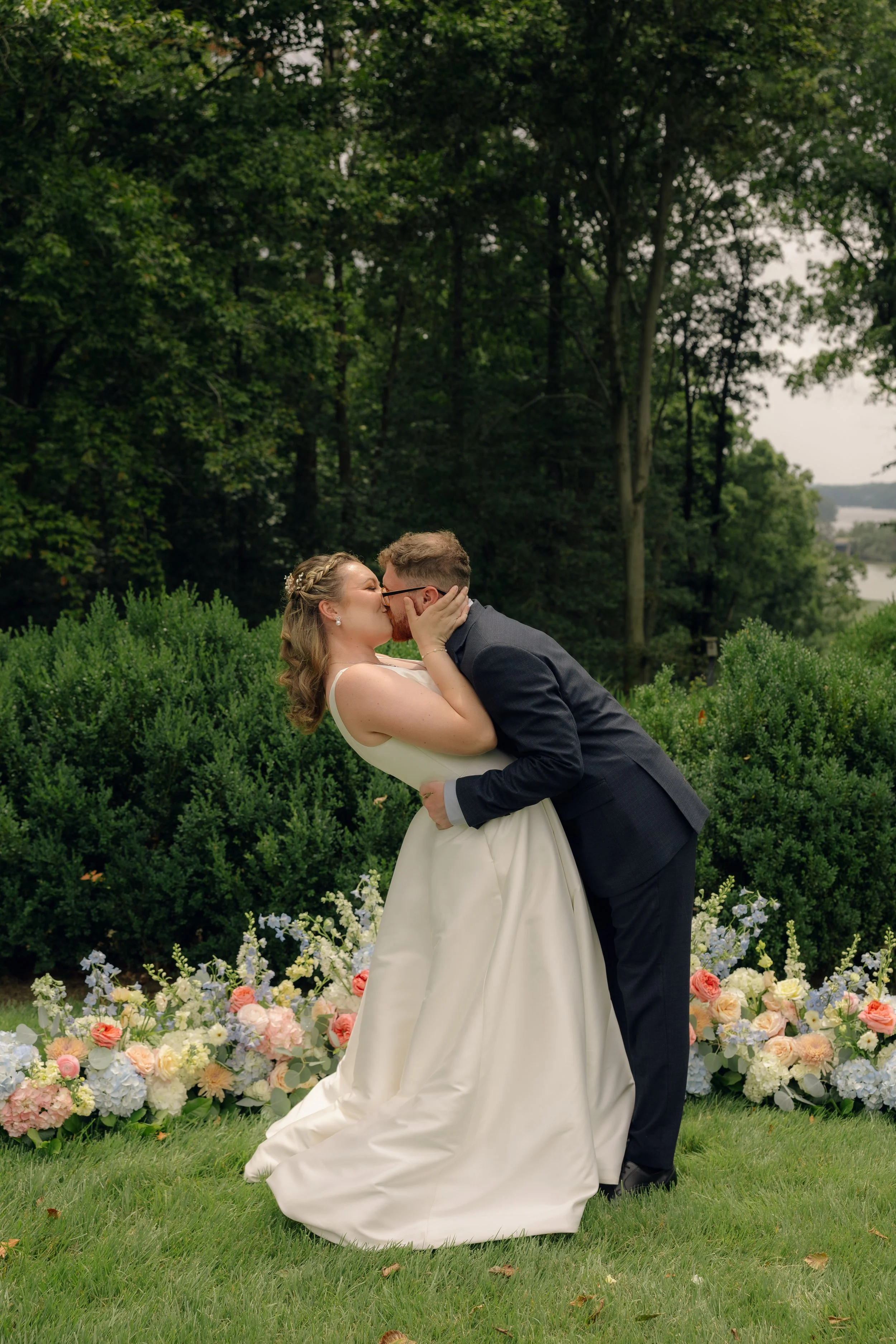 Couple sharing a kiss at their outdoor wedding ceremony, surrounded by flowers and greenery.