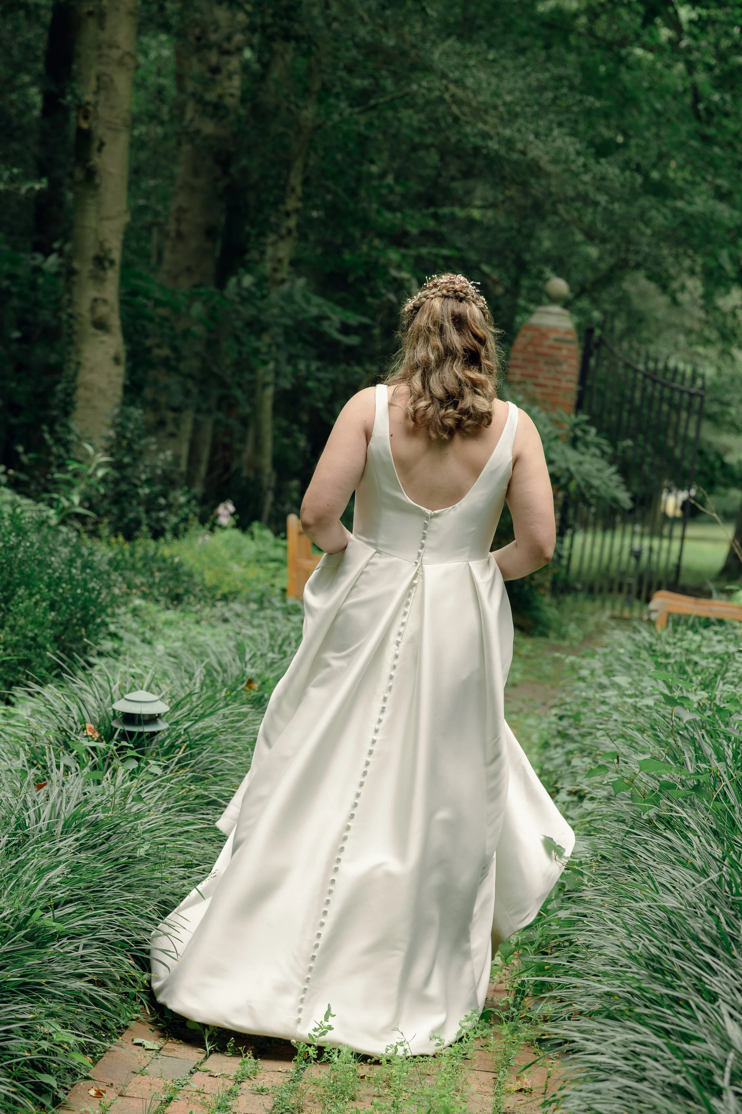 A woman in a white wedding dress standing in a garden with lush green foliage, trees, and a brick gate in the background.