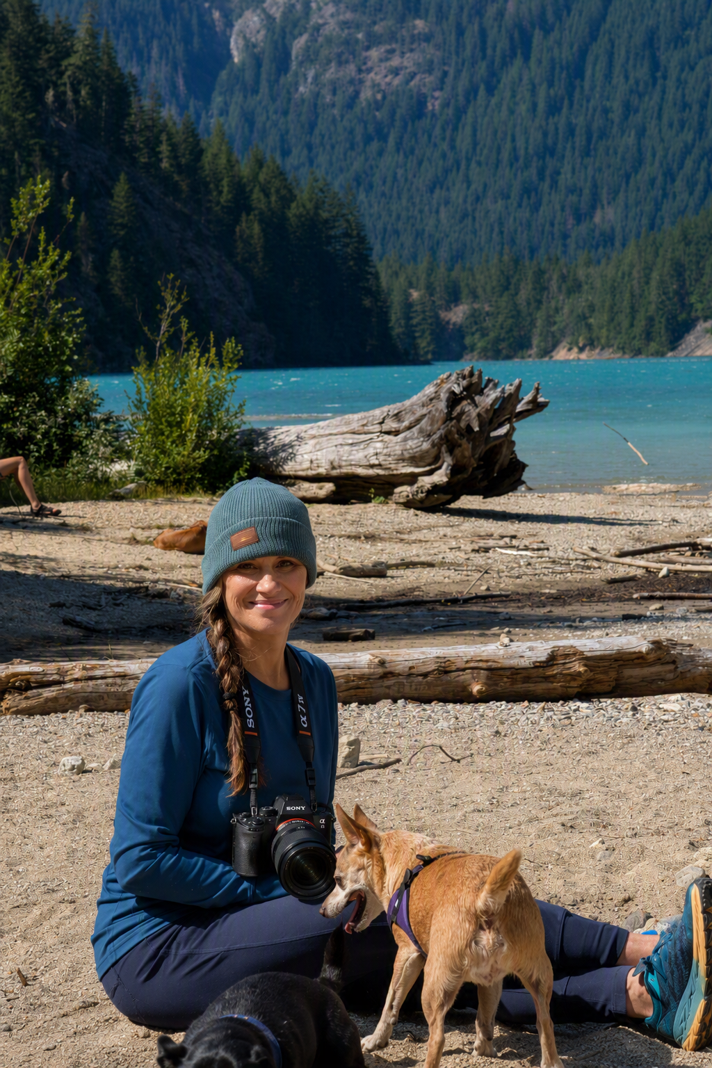 A woman smiling with a camera, sitting on a sandy beach near a turquoise lake surrounded by pine trees and mountains, with two dogs nearby.