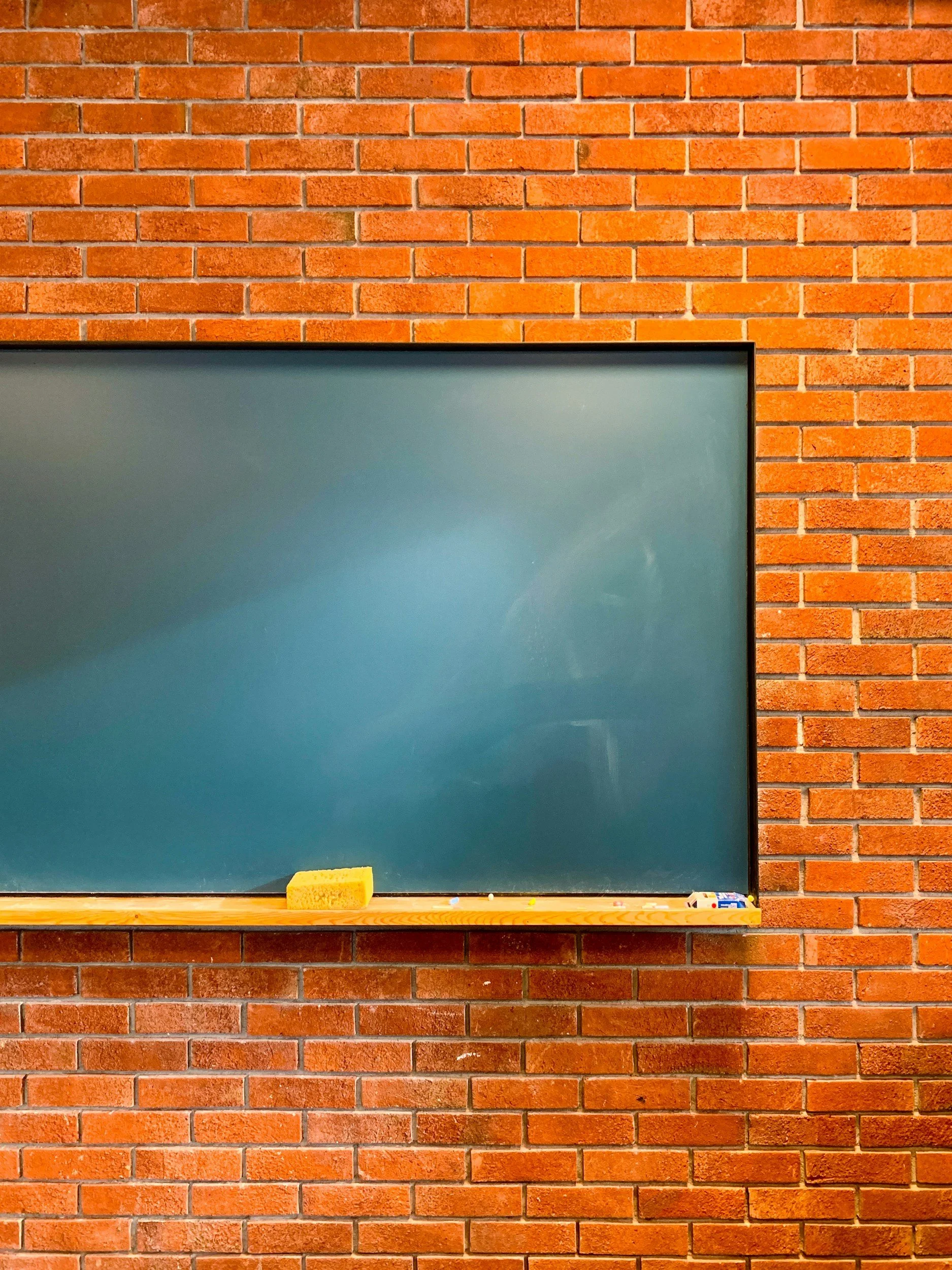 A chalkboard mounted on a red brick wall with a yellow sponge on the lower ledge of the chalkboard.