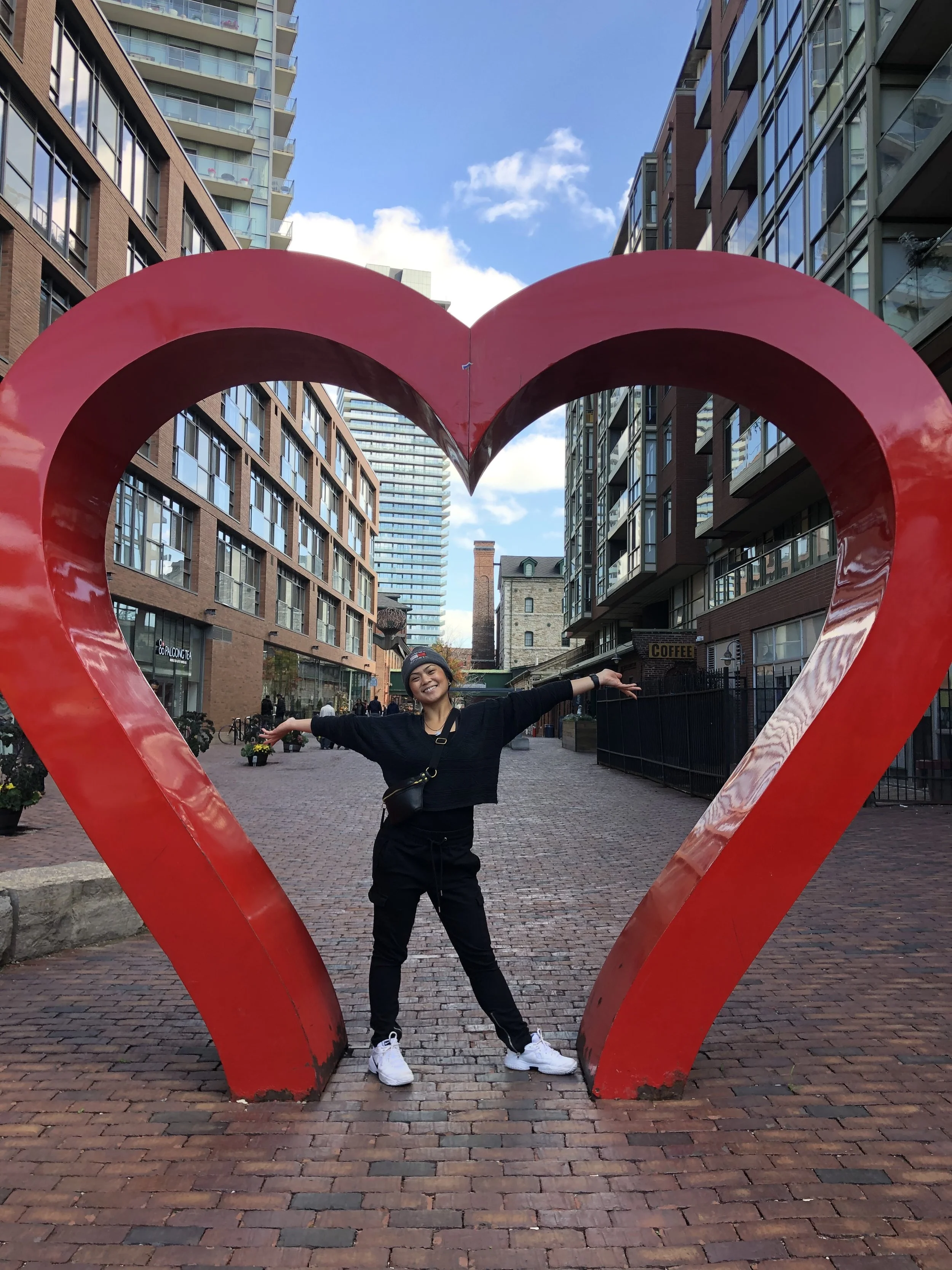A smiling woman standing with arms outstretched inside a large red heart-shaped sculpture on a city street with buildings in the background.