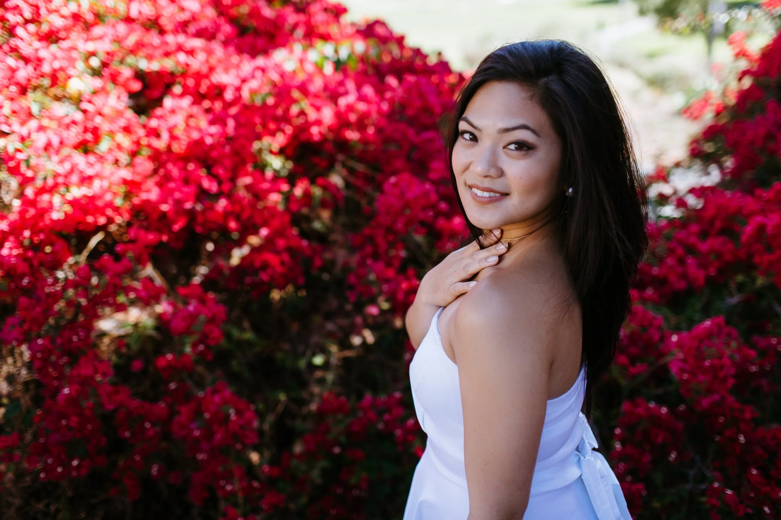 A woman with long dark hair smiling and looking at the camera, wearing a white dress, standing against a background of bright pink flowering bushes.