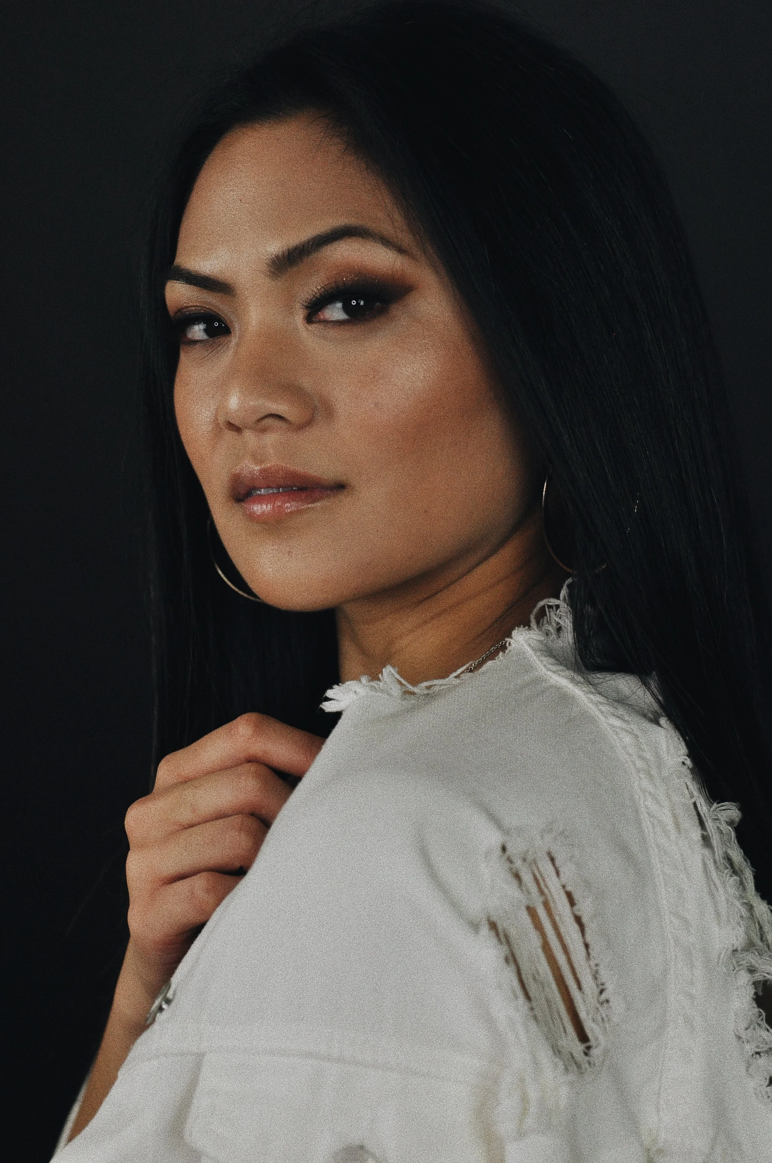 Close-up portrait of a woman with dark hair, makeup, and wearing hoop earrings, holding her shirt, against a dark background.