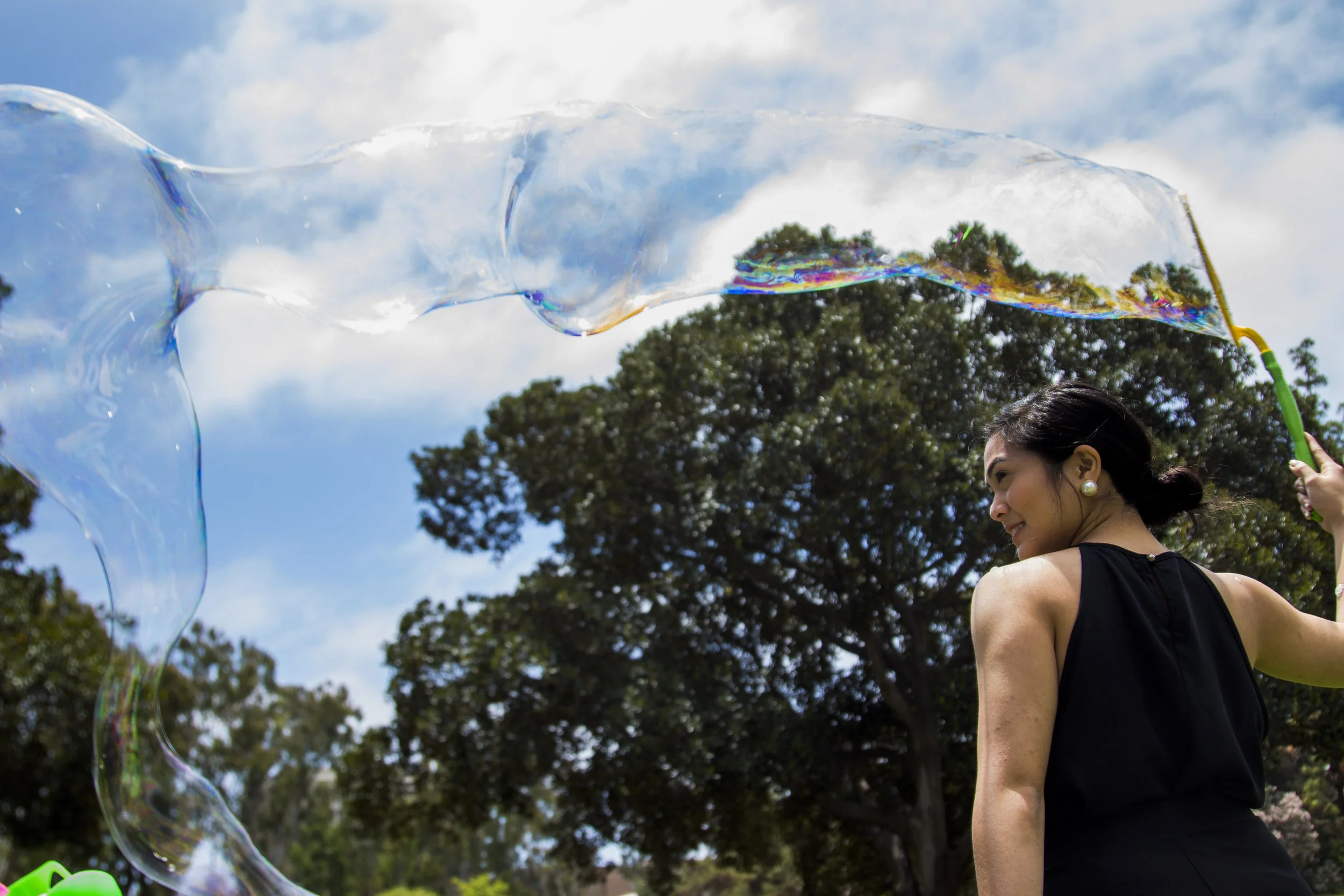 A woman in a black sleeveless dress playing with large soap bubbles outdoors on a sunny day, with trees and a blue sky with clouds in the background.