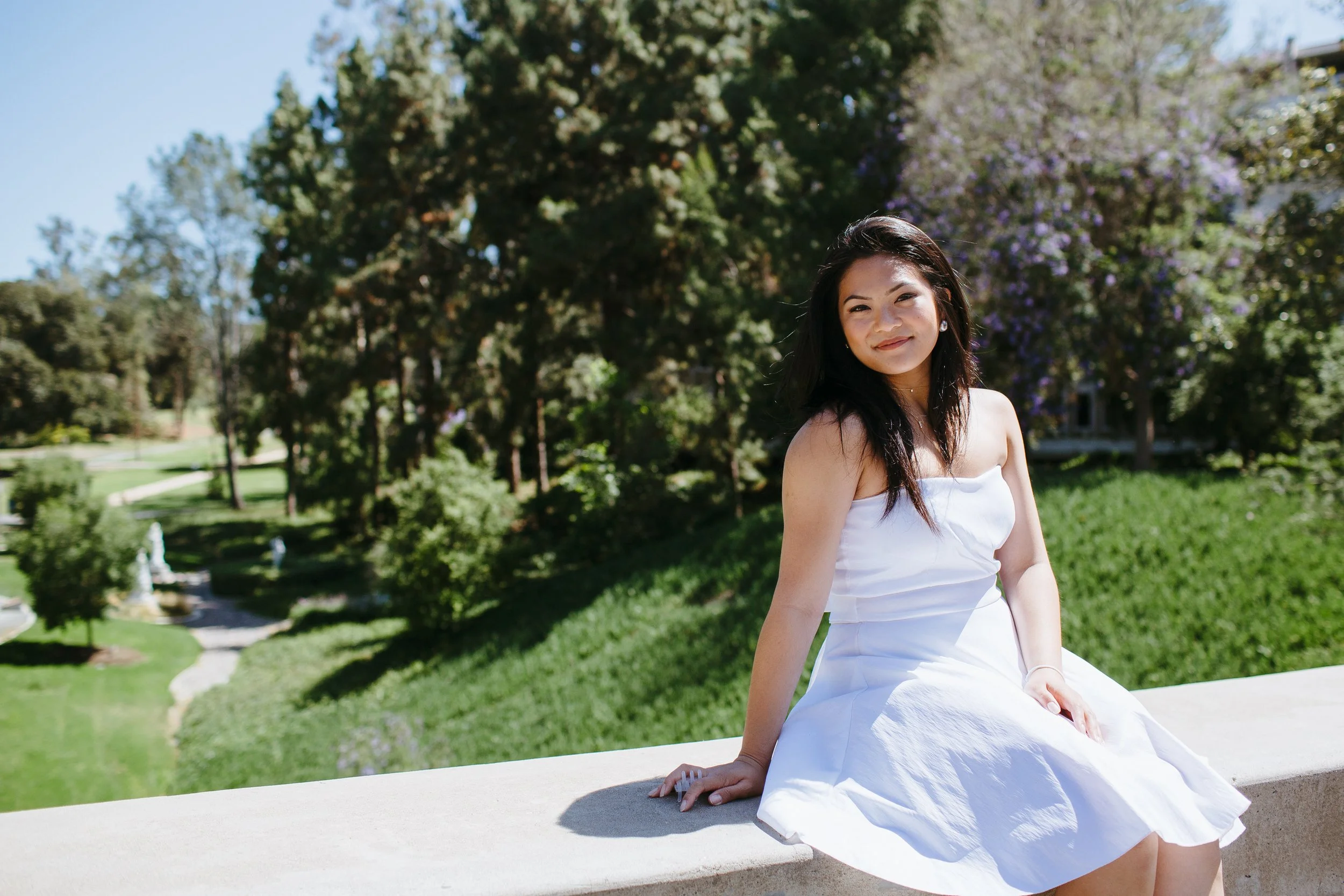 A woman in a white dress sitting outdoors on a concrete ledge, smiling with a background of trees and greenery on a sunny day.
