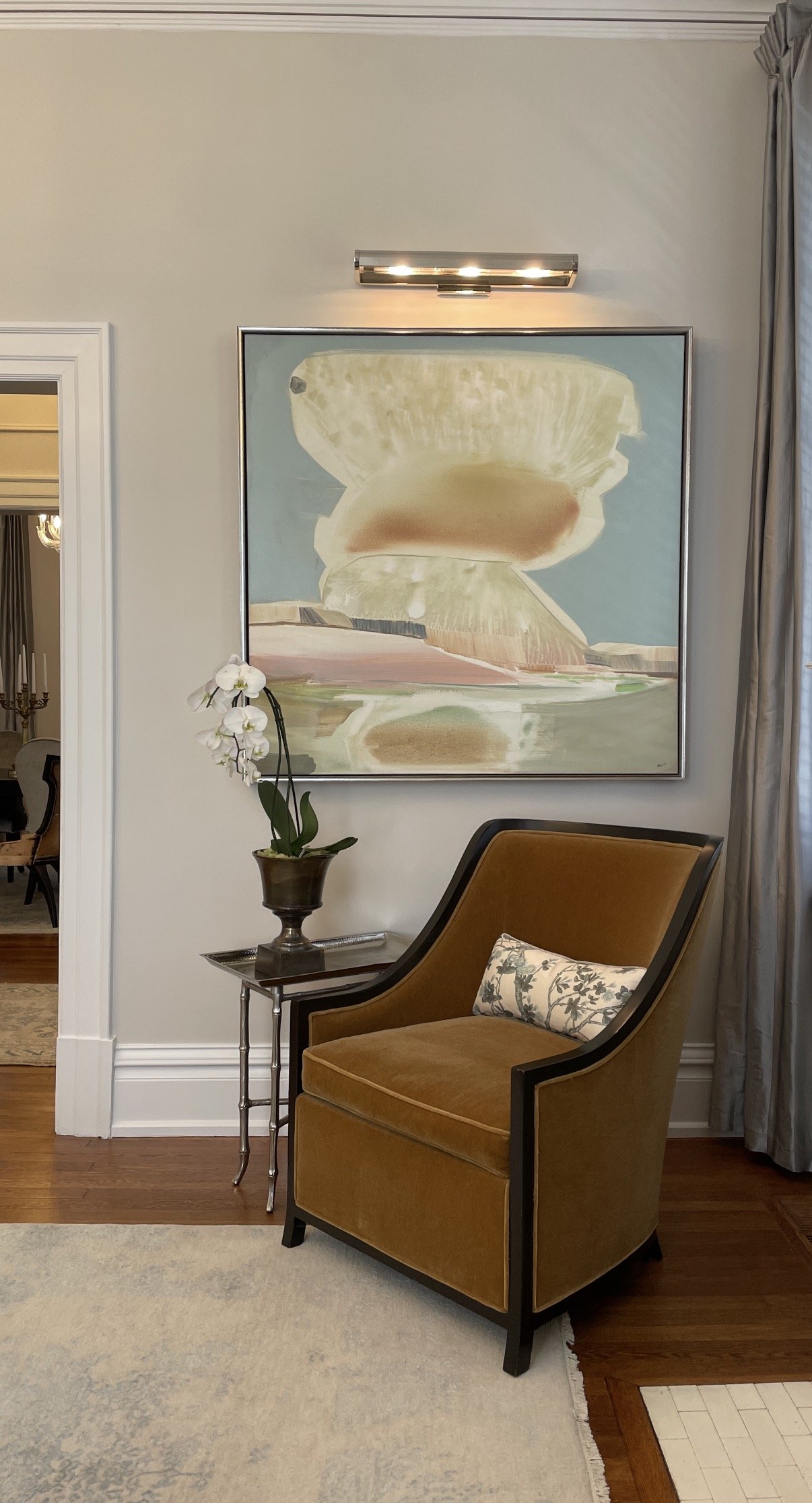 A room with a large abstract painting with relaxing colors hanging on the wall, above a designer armchair with a pillow on it. A side table with an orchid and a tray is next to the armchair. The room has wood flooring and is beautifully curated.