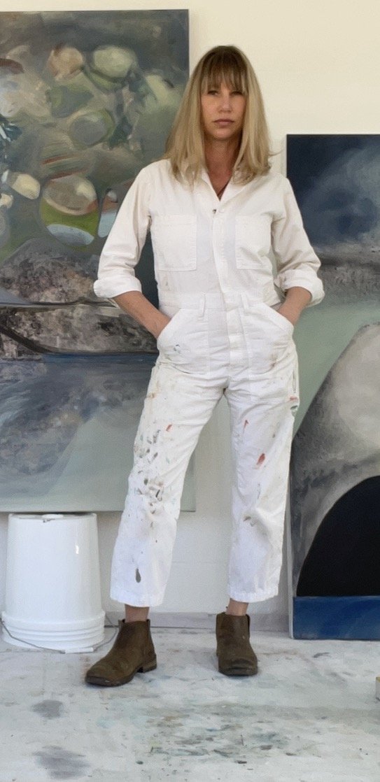 Woman, Shea Slemmer, with blonde hair wearing a white painter's jumpsuit and brown boots standing in an art studio with her paintings and an art bucket in the background.
