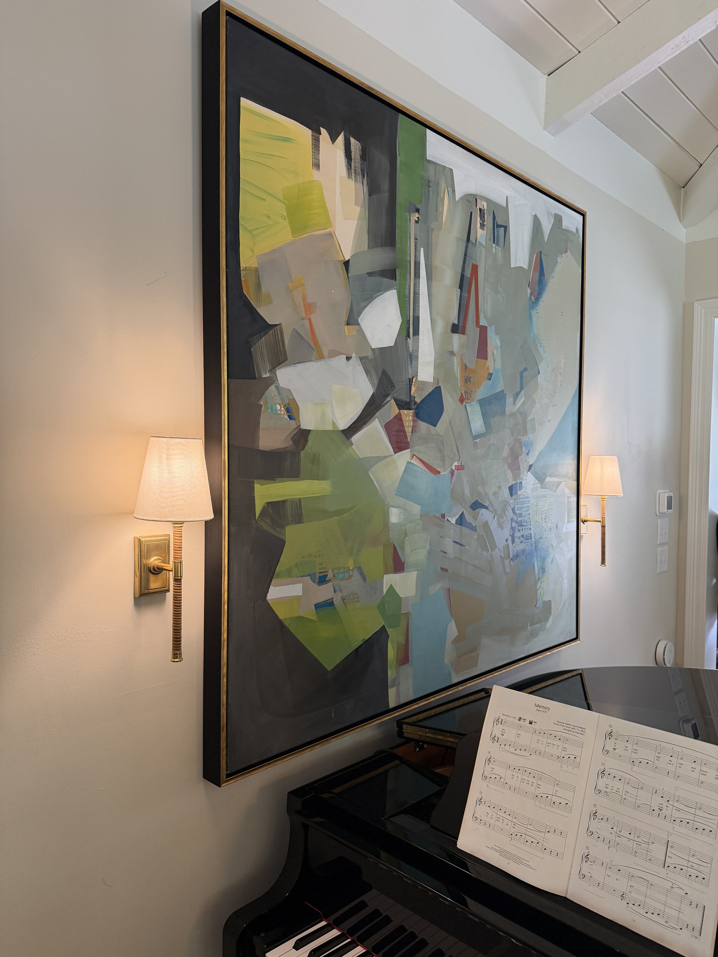 A large painting of a cityscape with buildings and trees, framed and hung on a wall. Two wall-mounted sconces with lamps flank the painting. Below, a black grand piano with open sheet music on its music stand, adjacent to a dark-colored bench.