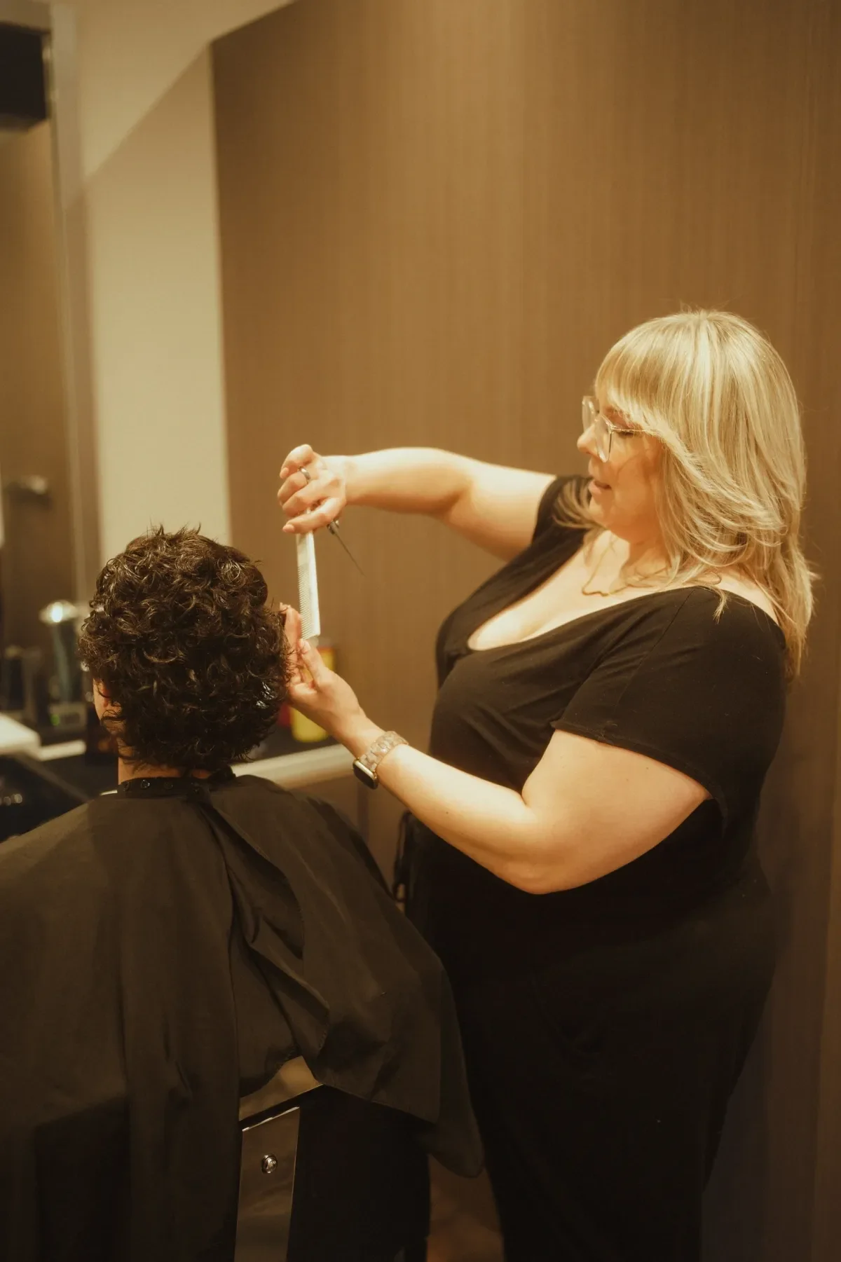 A makeup artist applying makeup to a woman with short curly hair, sitting in a salon or makeup studio.