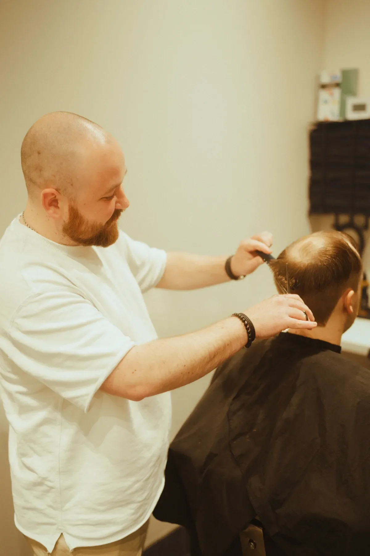 A bald man with a red beard cutting another person's hair in a salon. The client has short, light brown hair and is wearing a black cape. The barber is wearing a white t-shirt and black beaded bracelets.