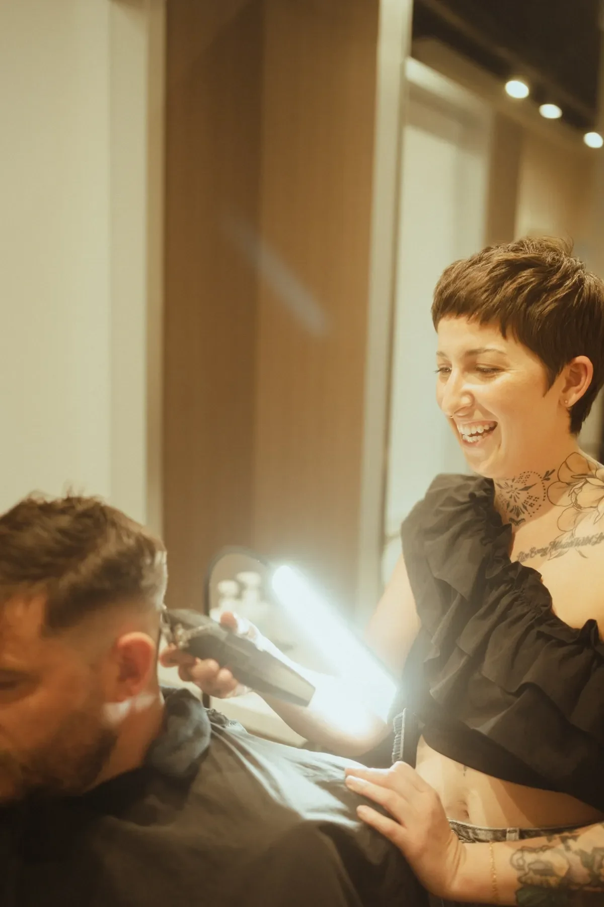 A woman with short brown hair and tattoos on her neck and arms laughs while giving a tattoo to a man in a tattoo parlor. The man is seated, wearing glasses, and the woman appears to be holding a tattoo gun.